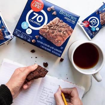 Someone writing in a journal while holding a Fiber One brownie. Nearby on the table sits a cup of coffee and a box of Fiber One 70 calorie brownies. - Link to social post