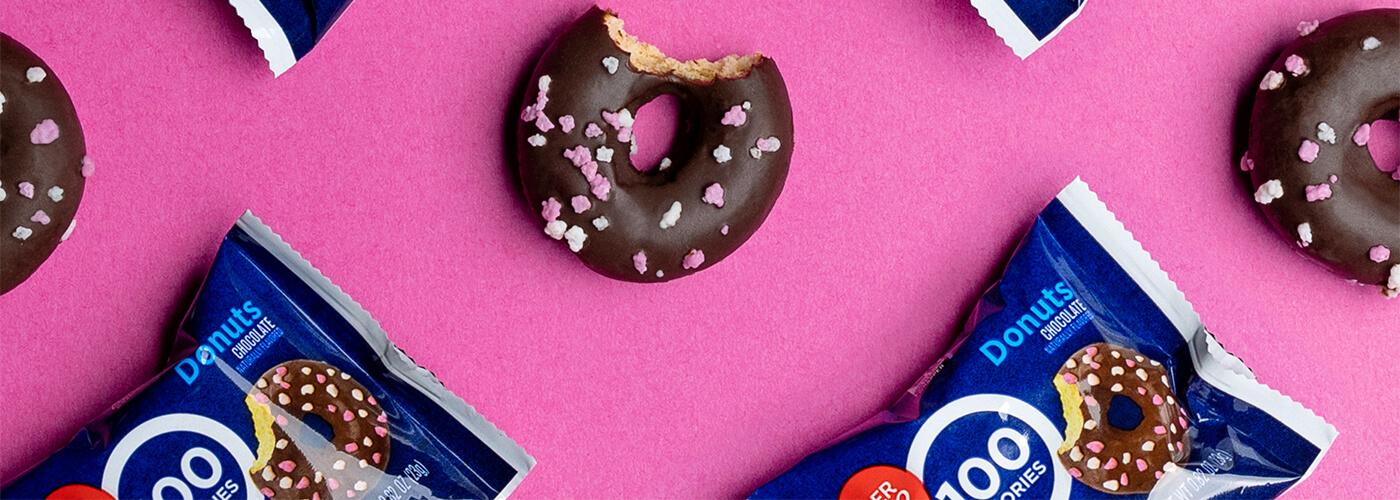 Packaged chocolate frosted donuts with sprinkles and a bitten donut, displayed on a pink background.