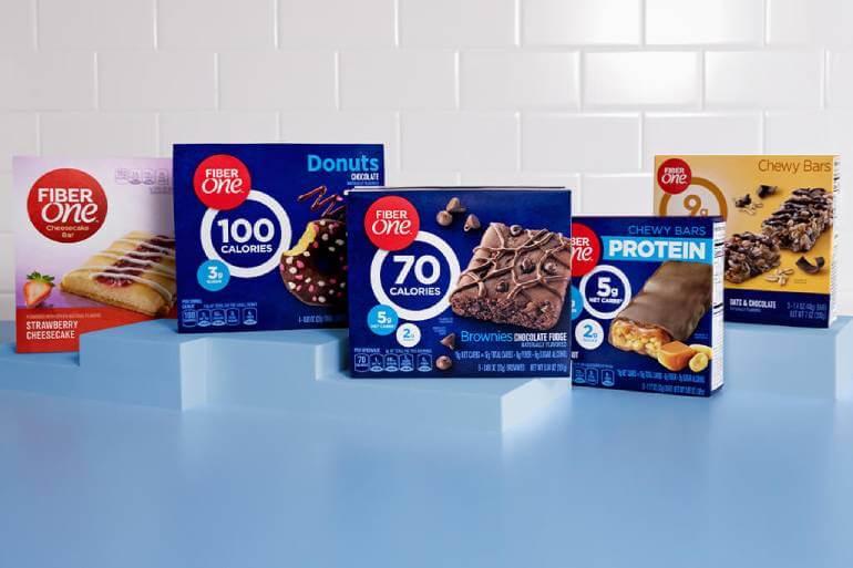 A display of five varieties of Fiber One snack boxes, including Strawberry Cheesecake Bars, Mini Donuts, Brownies, and Chewy Bars with Protein, arranged on a light blue surface against a tiled background